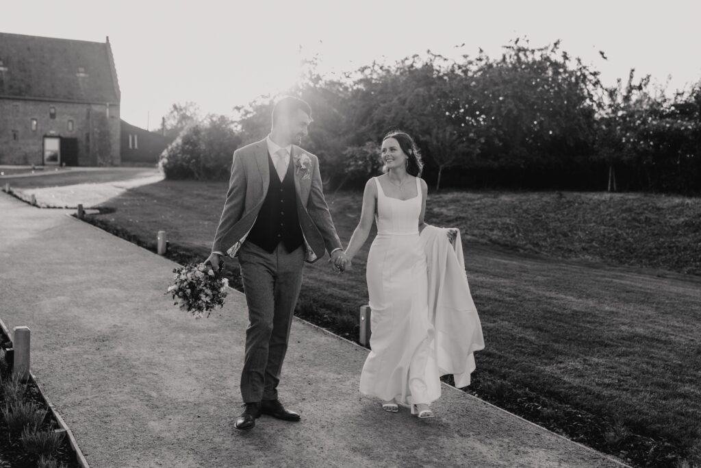 Bride and groom walking hand in hand through fields at Copdock Hall during golden hour