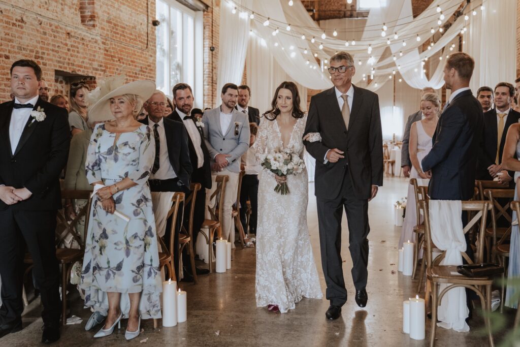 Bride walking down the aisle at Godwick Great Barn as groom wipes tears