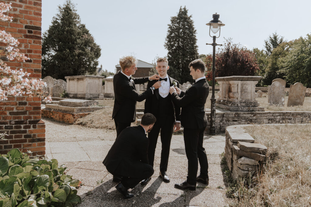 Groom adjusting his tie outside the church with groomsmen laughing in the background