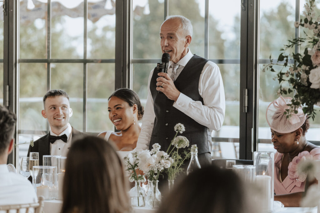 Father of the bride raising a glass during his heartfelt wedding speech in the orangery at High House