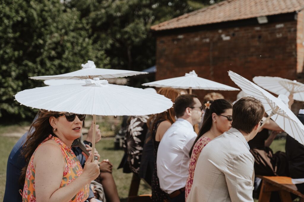 Guests smiling under parasols during hot June wedding ceremony in Suffolk