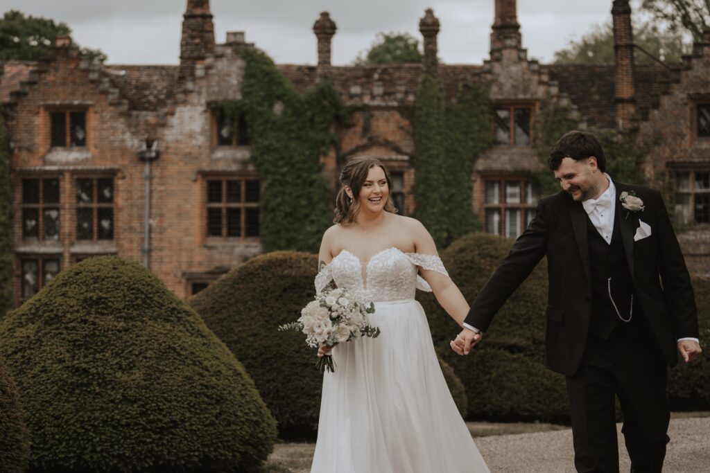 Bride and groom walking hand-in-hand across Seckford Hall’s front lawn