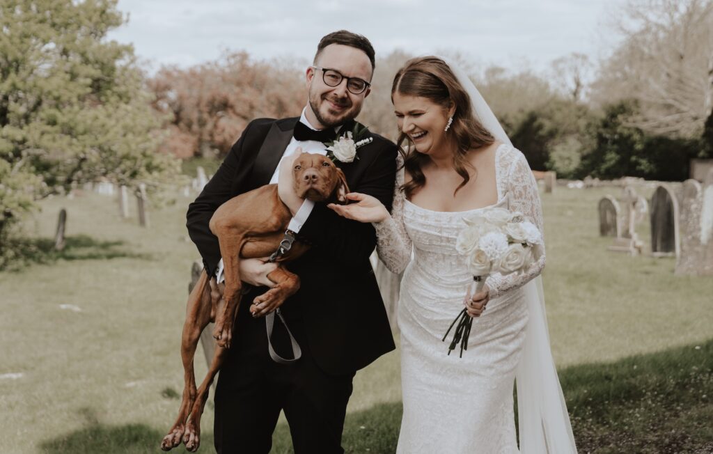 A bride and groom laughing and holding their dog on their wedding day in Kent